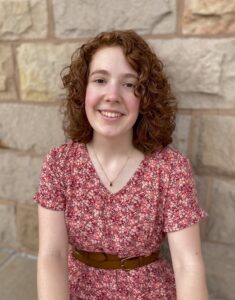 Jacey Samic headshot - woman standing against a wall and smiling