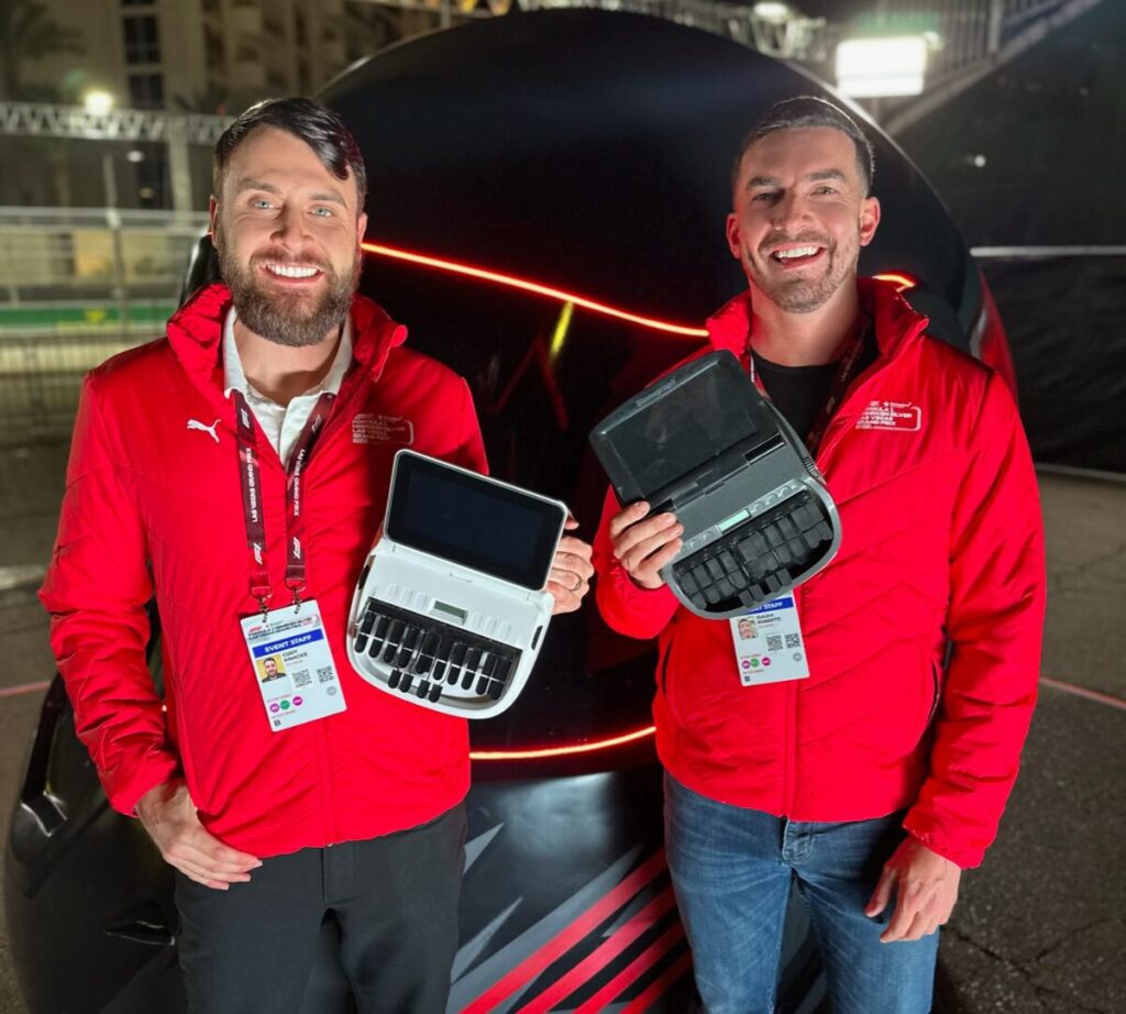 Cody Knacke (l) and Isaiah Roberts (r) at a recent Formula One racing event. They're holding steno machines in front of an oversized helmet