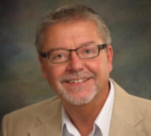 Head shot of Bill Sammons in a beige jacket, white collared shirt, short cropped hair and glasses.